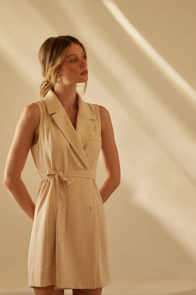 Woman wearing a beige sleeveless blazer-style mini dress with a waist tie and lapel collar.