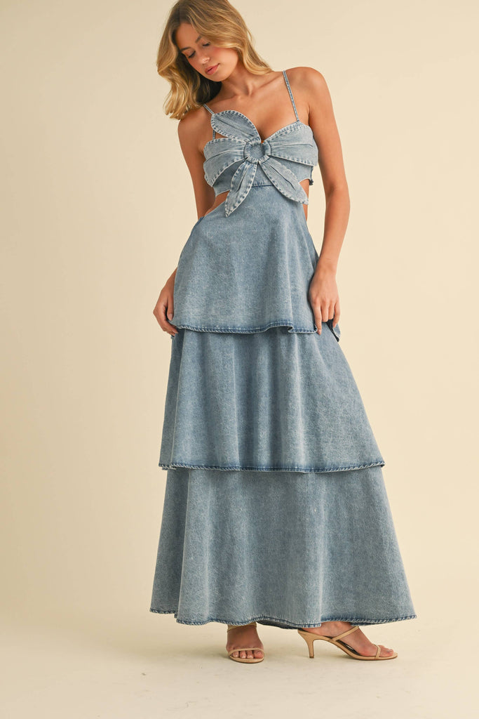 Woman wearing a denim maxi dress with a large front bow, side cutouts, spaghetti straps, and tiered skirt.