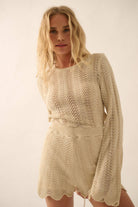 Woman wearing a beige knit romper with bell sleeves, scalloped hems, and open-weave detailing.
