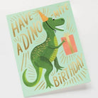 A birthday card featuring a green dinosaur wearing a party hat and holding a gift, with the text "Have a Dino-Mite Birthday," paired with a gold envelope.