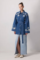 Model wearing a belted denim jacket with white floral embroidery and wide sleeves