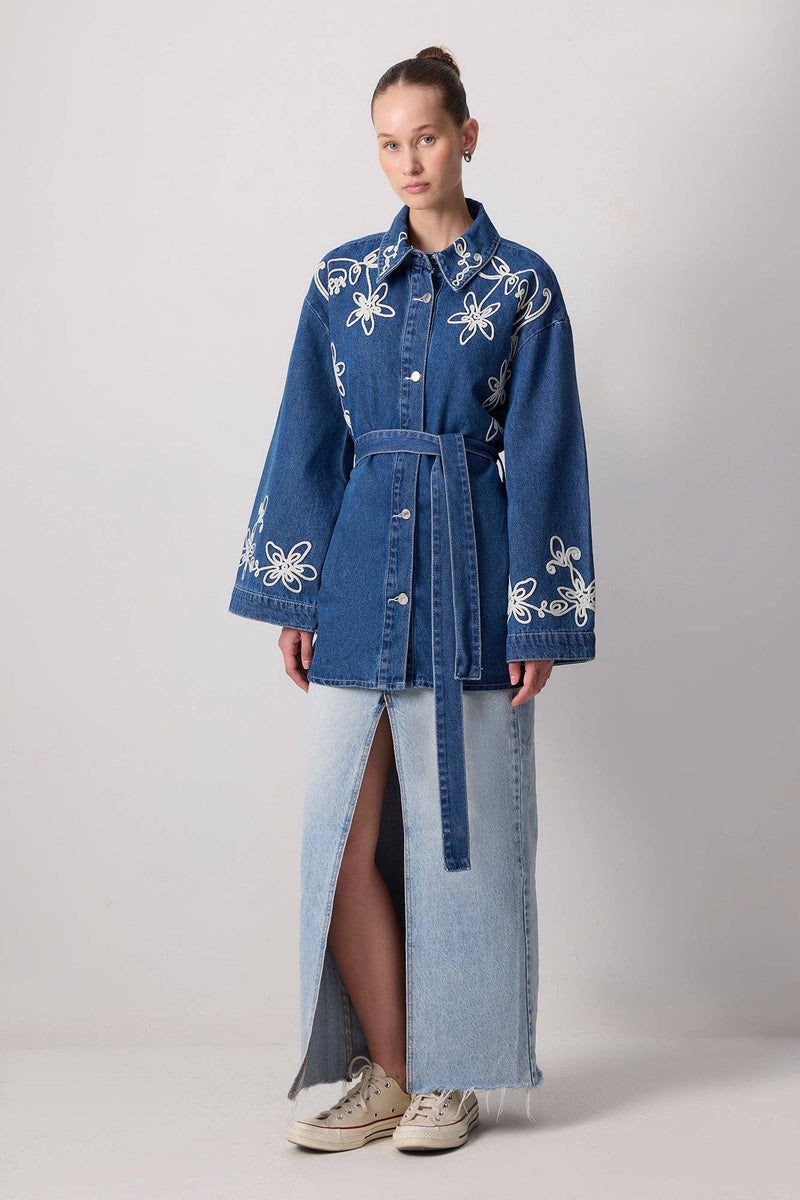Model wearing a belted denim jacket with white floral embroidery and wide sleeves