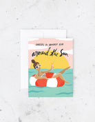 Birthday card with an illustration of a woman in a floatie holding a drink with the words “Cheers to another year around the sun”.