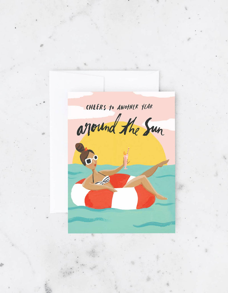 Birthday card with an illustration of a woman in a floatie holding a drink with the words “Cheers to another year around the sun”.