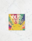 Greeting card with a smiling sun, rainbow-colored rays, and gold foil text that reads “You Are My Sunshine.”