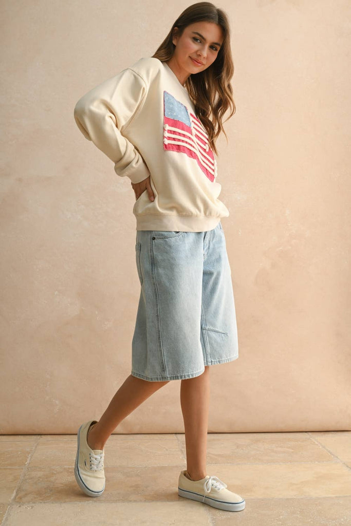 Woman wearing a cream sweater with a textured rope-stripe American flag design, paired with light wash jeans.