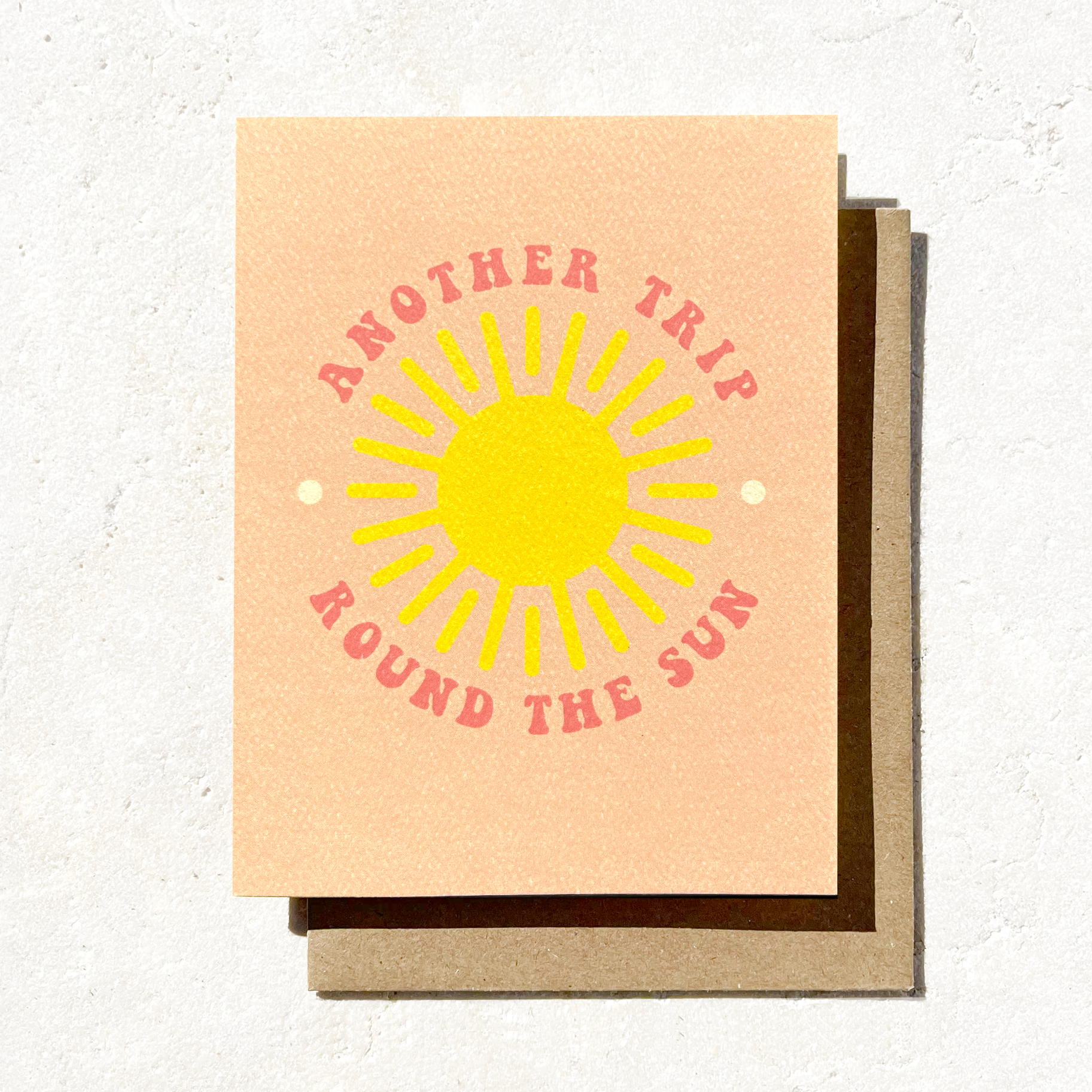 A greeting card with a vibrant yellow sun design surrounded by the phrase "Another Trip Round the Sun" in pink, set against a peach background.