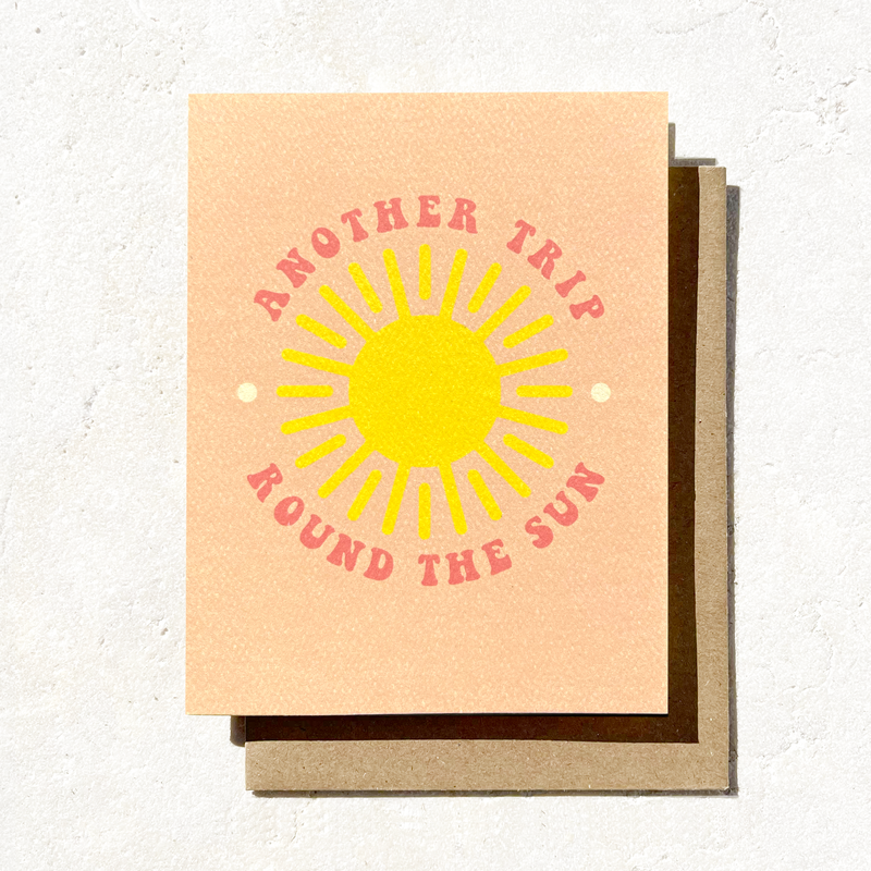 A greeting card with a vibrant yellow sun design surrounded by the phrase "Another Trip Round the Sun" in pink, set against a peach background.