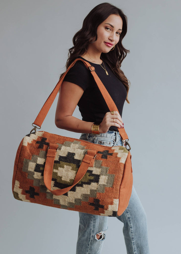 Earth-tone woven weekender bag with geometric pattern in black, green, and cream, held by woman in jeans and black top