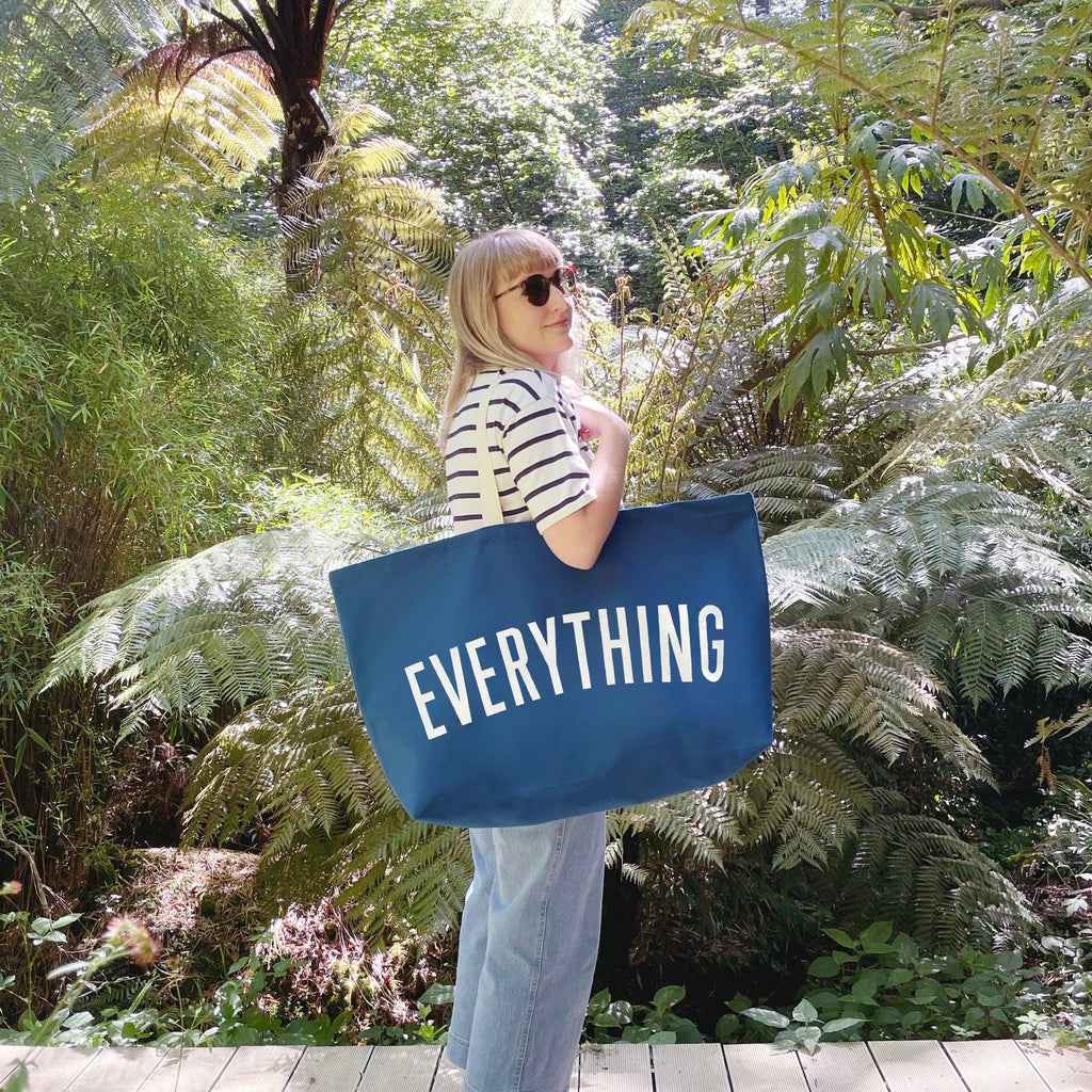 Oversized navy blue tote bag with cream handles and bold white “EVERYTHING” text, held by a hand against a light wood background