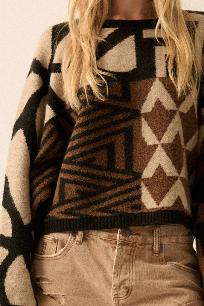 Model wearing a patterned knit sweater with bold geometric designs in neutral and contrasting tones, styled with casual bottoms.