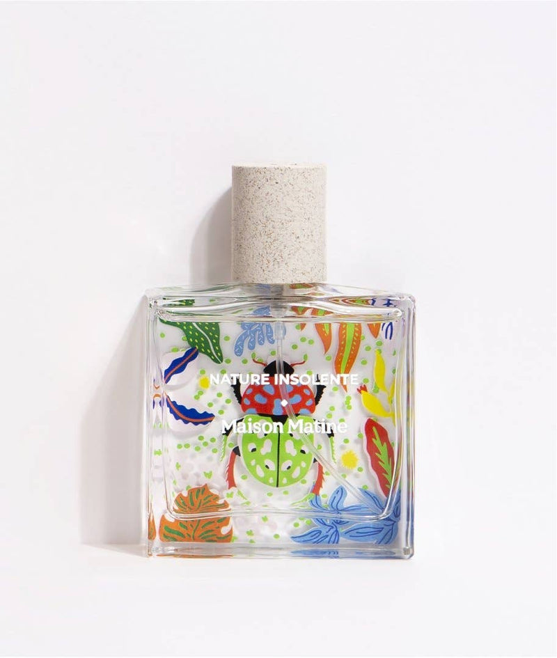 Maison Matine Nature Insolente perfume bottle with colorful leaf and beetle illustrations, placed on a vibrant yellow fabric and surrounded by green foliage.