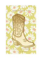A brass bookmark shaped like a cowgirl boot with floral engravings, displayed on a daisy-patterned yellow backing card.
