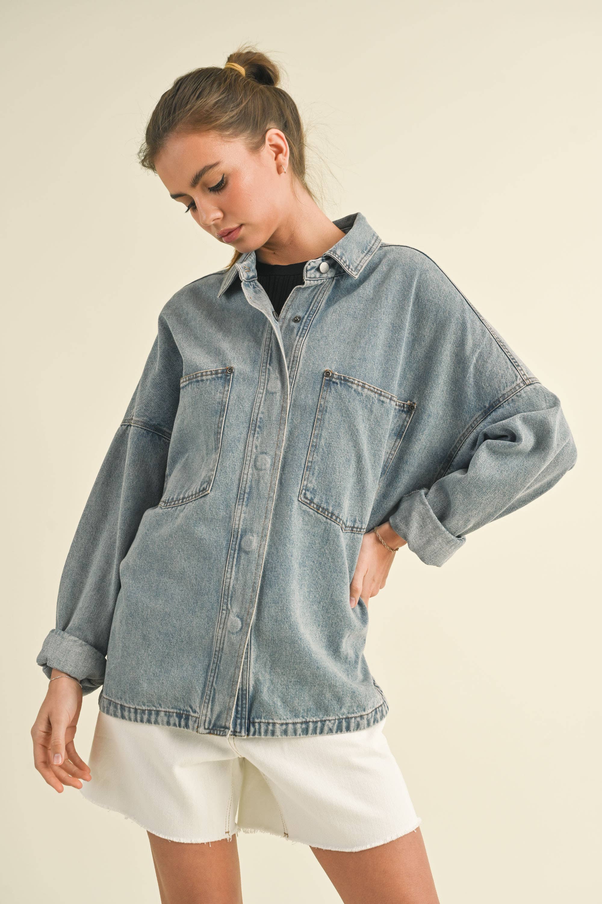 Model in an oversized denim jacket with front pockets. First image: jacket buttoned, paired with white shorts. Second image: jacket open, over a grey tank and denim pants.