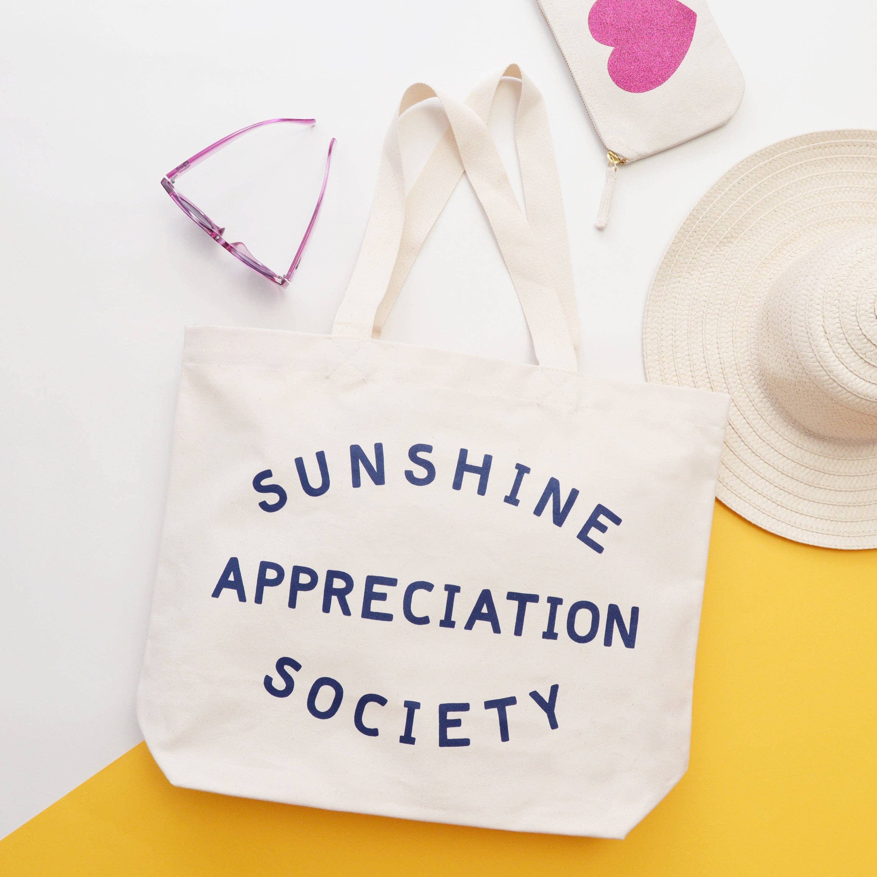 Cream canvas tote bag with “Sunshine Appreciation Society” printed in navy, filled with flip-flops, sunglasses, and travel essentials on a pink background