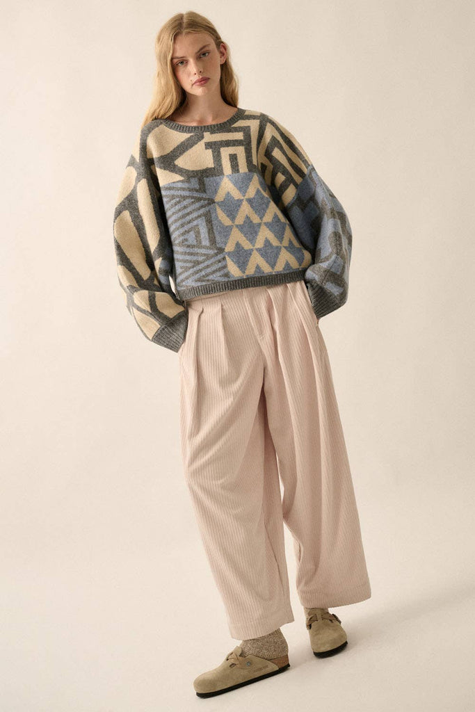 Model wearing a patterned knit sweater with bold geometric designs in neutral and contrasting tones, styled with casual bottoms.