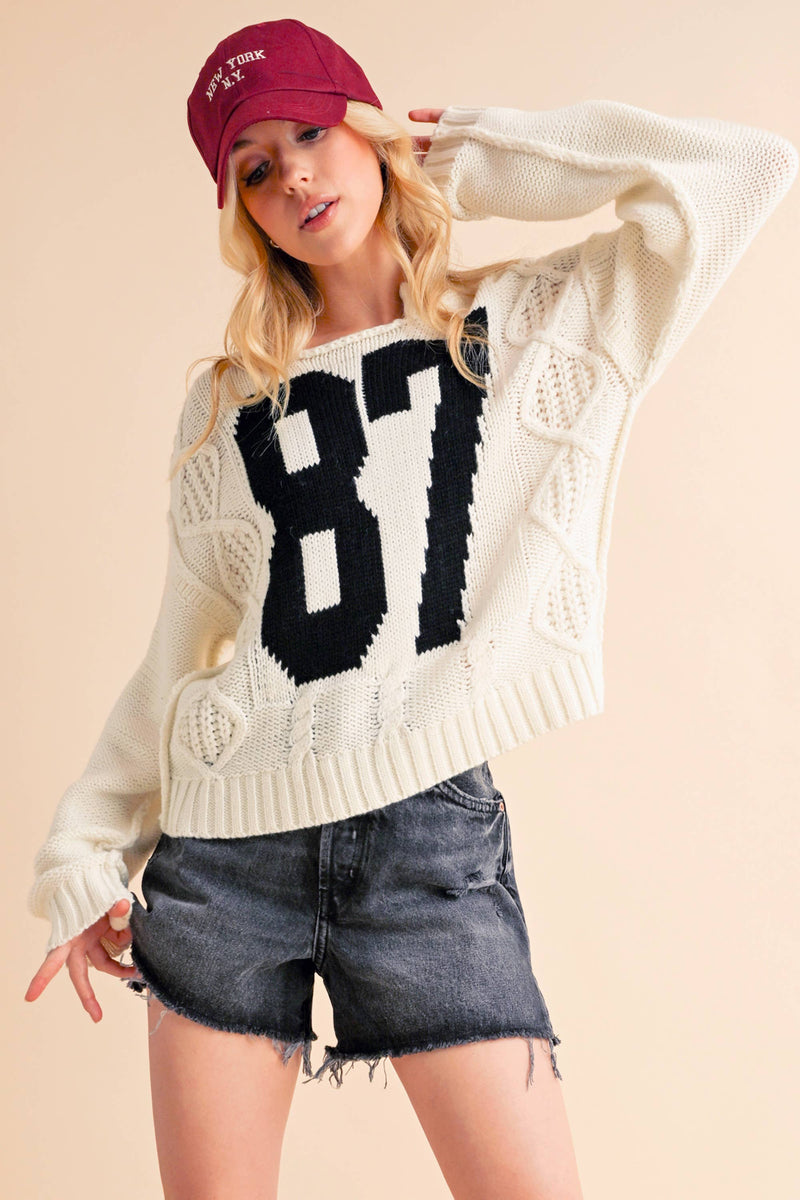 Woman wearing a cream knit sweater with a bold black "87" graphic and dark denim shorts.