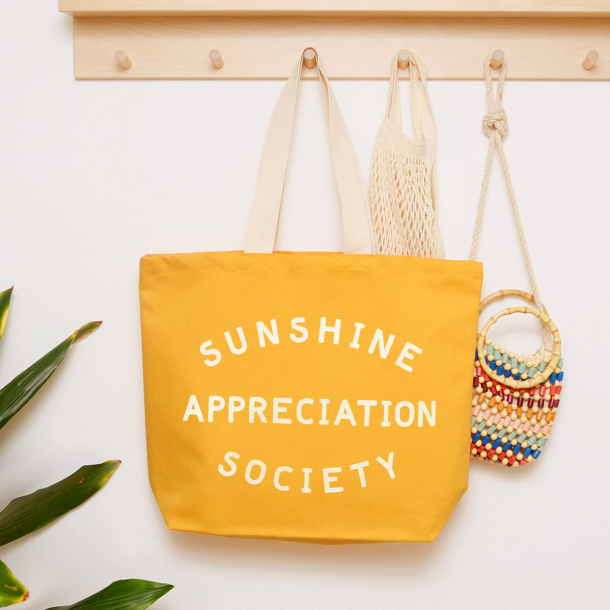 Yellow canvas tote bag with “Sunshine Appreciation Society” printed in white, held up by a hand in front of a sage green wall