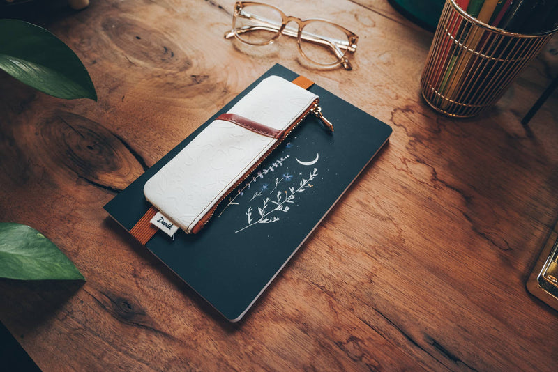 A white vegan leather notebook pouch with an embossed floral design, tan accents, and a gold zipper, attached to its packaging.