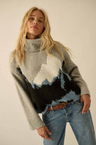 Person wearing a gray sweater with mountain design and blue jeans on a plain background