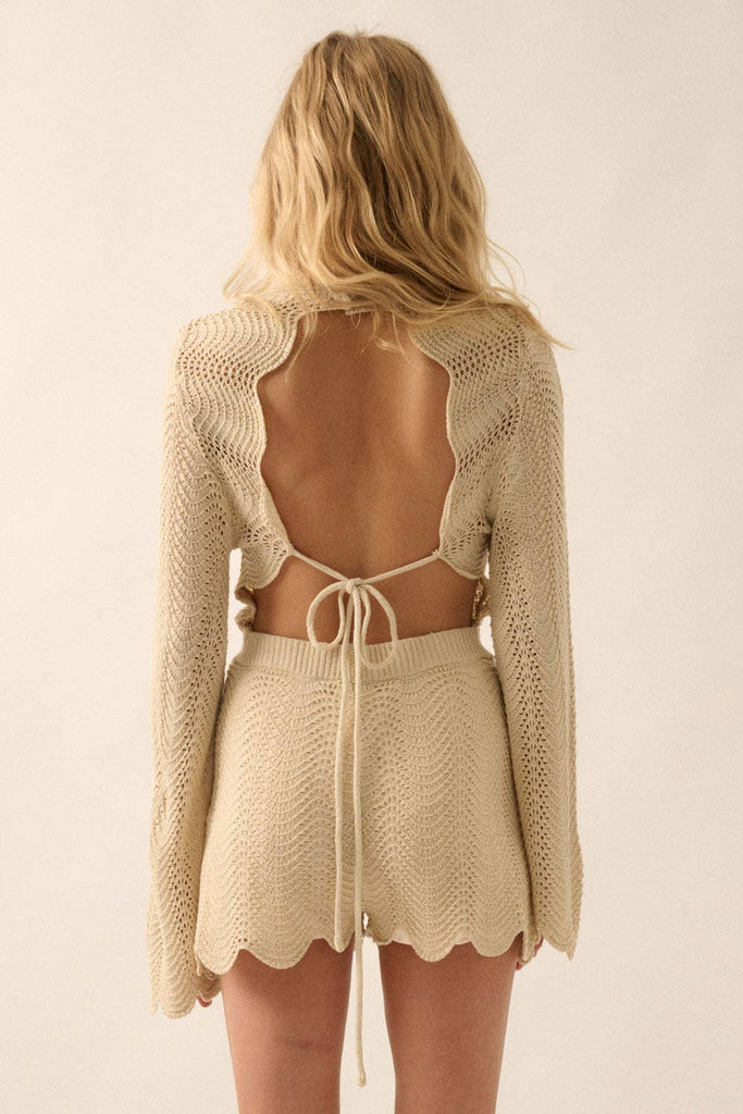Woman wearing a beige knit romper with bell sleeves, scalloped hems, and open-weave detailing.