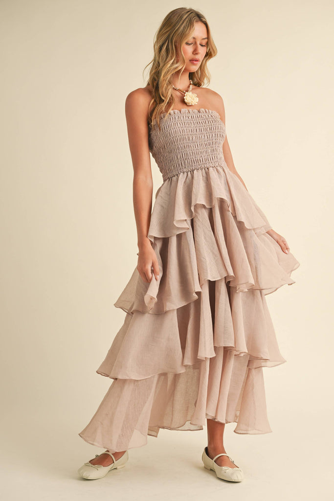 Woman wearing a strapless ruffled maxi dress with a smocked bodice, available in taupe and black.
