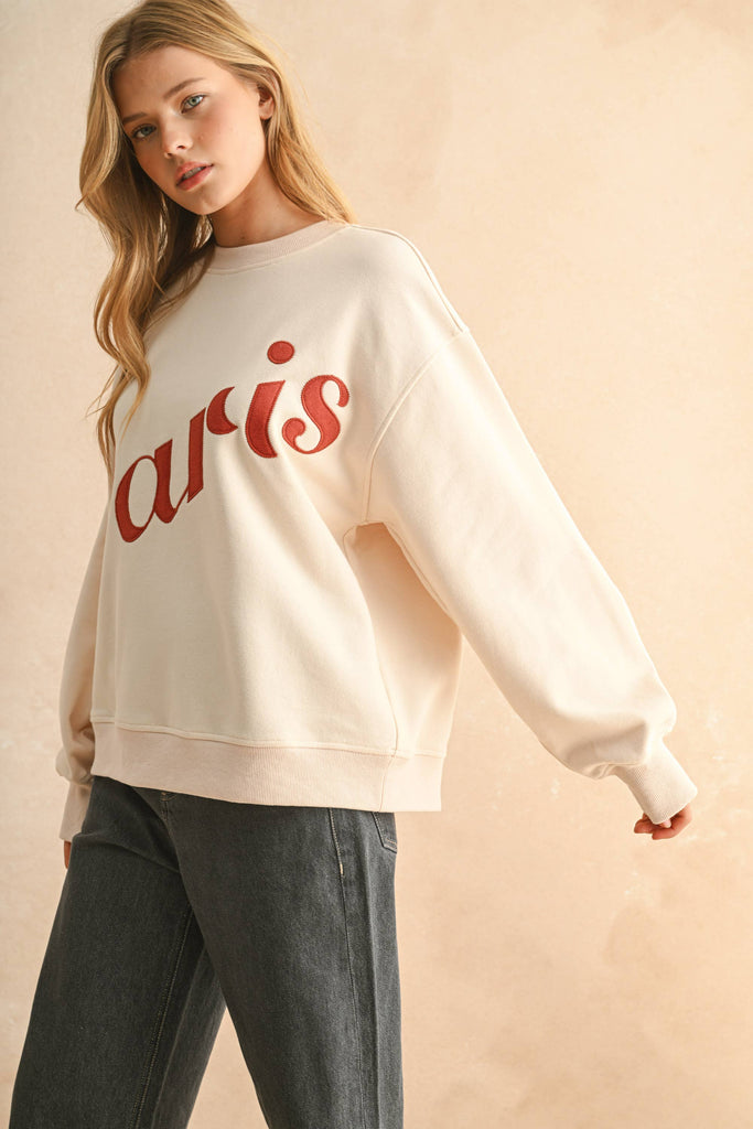 Woman wearing a cream sweater with bold red embroidered “Paris” lettering, paired with dark denim jeans.