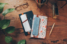 A white vegan leather notebook pouch with an embossed floral design, tan accents, and a gold zipper, attached to its packaging.