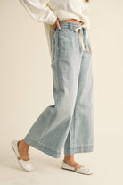 Woman wearing wide-leg light denim pants with a tie-front waist, paired with a white blouse and ballet flats.