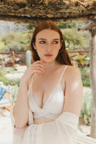 Model wearing a scalloped-edge lace bralette in burgundy and blue tones, styled with layered necklaces and velvet pants.