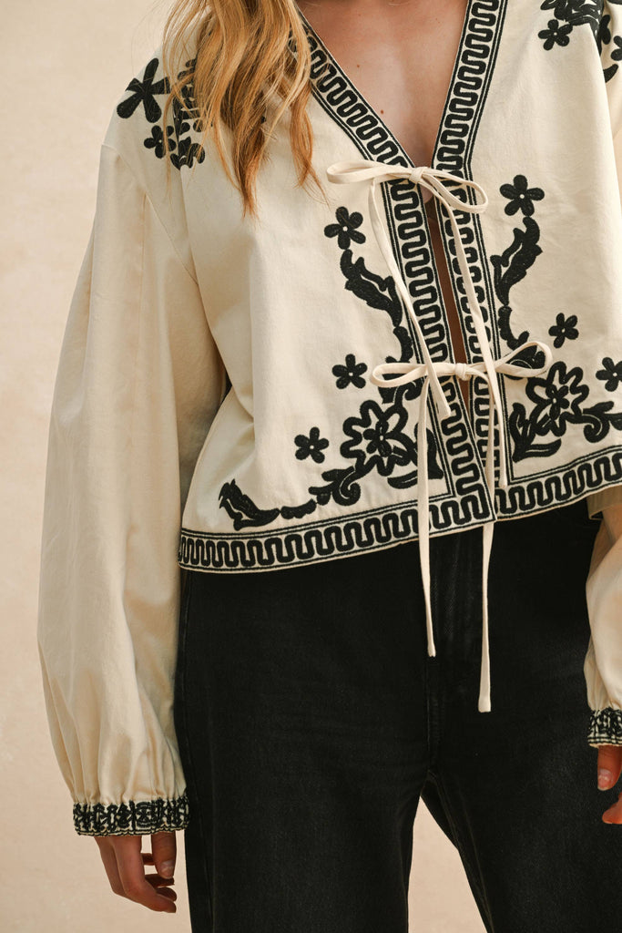 Woman wearing cream blouse with black floral embroidery, lace-up front, and long billowy sleeves.