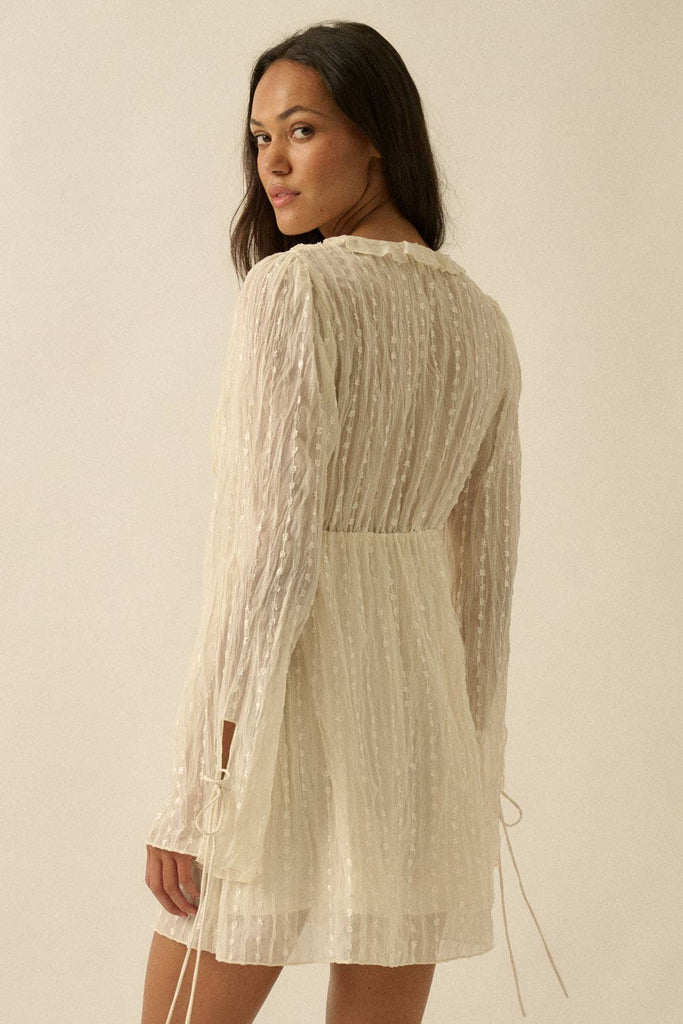 Woman wearing a cream mini dress with sheer long sleeves, button front, and tie details.
