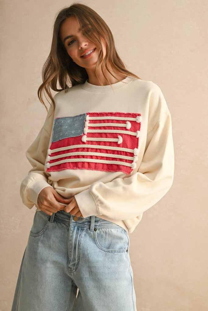 Woman wearing a cream sweater with a textured rope-stripe American flag design, paired with light wash jeans.