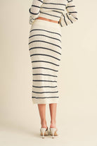 Woman wearing a high-waisted, ivory and black striped knit midi skirt with slide sandals.