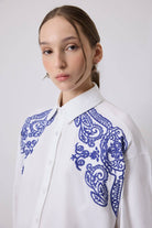 Woman wearing a white oversized shirt with bold blue embroidery on shoulders