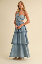 Woman wearing a denim maxi dress with a large front bow, side cutouts, spaghetti straps, and tiered skirt.
