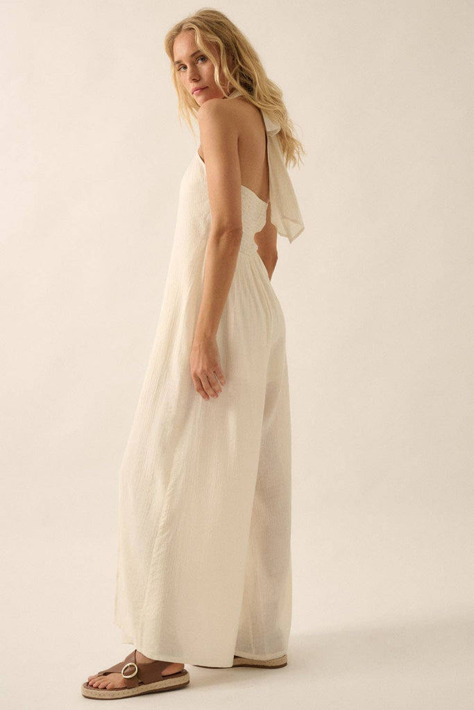 Woman wearing a white halter jumpsuit with tie-back neckline and wide-leg pants, styled with sandals.