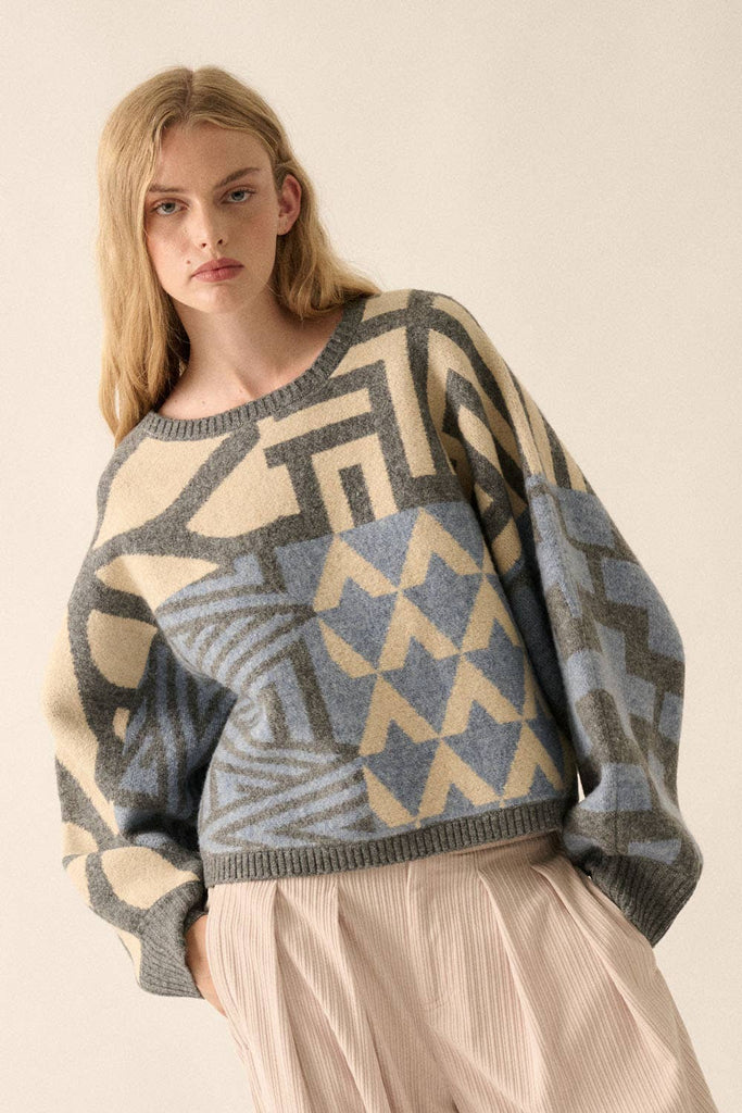 Model wearing a patterned knit sweater with bold geometric designs in neutral and contrasting tones, styled with casual bottoms.