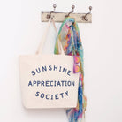 Cream canvas tote bag with “Sunshine Appreciation Society” printed in navy, filled with flip-flops, sunglasses, and travel essentials on a pink background