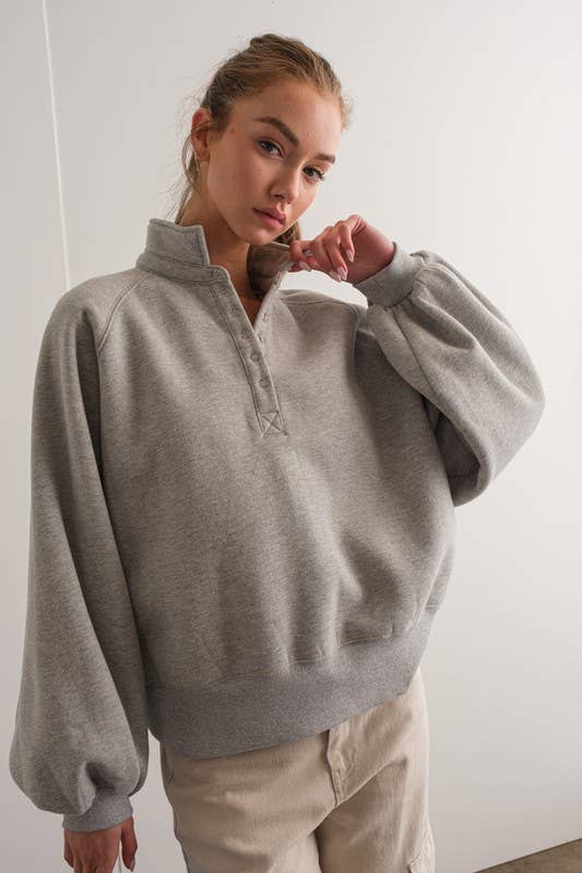 Woman wearing an oversized gray pullover sweatshirt with quarter snaps and voluminous sleeves, paired with beige pants.