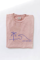Folded soft pink-colored t-shirt with a blue minimalist palm tree, wave, and "LIVE LOVE SURF" graphic.