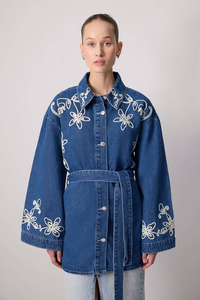 Model wearing a belted denim jacket with white floral embroidery and wide sleeves