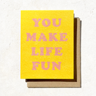 A yellow greeting card with pink text that reads "You Make Life Fun," paired with a brown envelope on a textured surface.