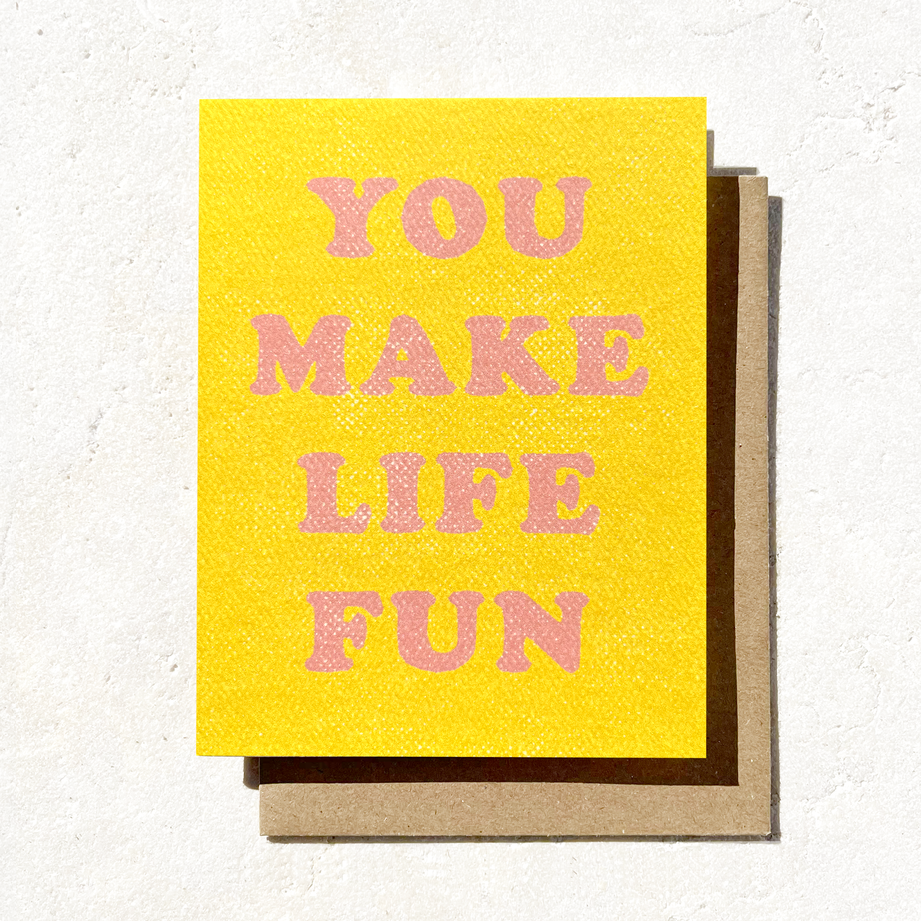 A yellow greeting card with pink text that reads "You Make Life Fun," paired with a brown envelope on a textured surface.