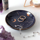 A round ceramic jewelry dish with a celestial design, including golden moons, stars, and constellations on a dark background.