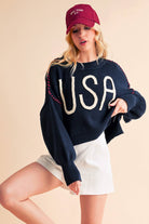 A woman wearing a navy sweater with the text 'USA' in white letters, paired with white shorts and a red cap.