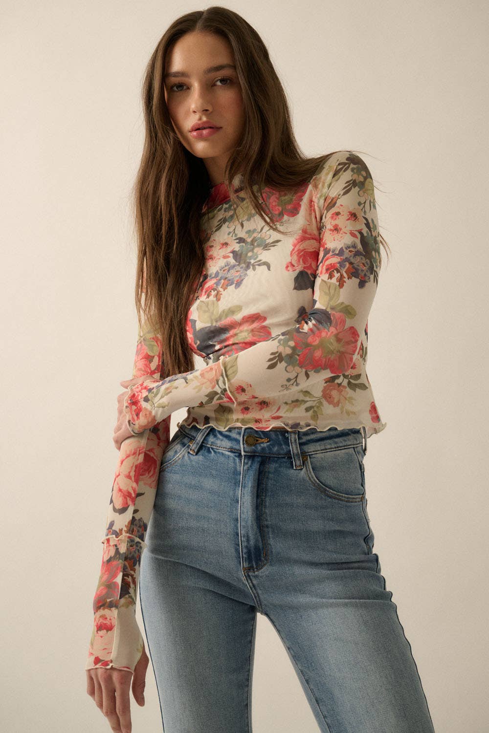 Model wearing a fitted sheer mesh long sleeve top with red, pink, and green floral print, paired with blue jeans.