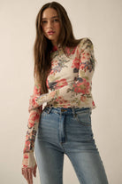 Model wearing a fitted sheer mesh long sleeve top with red, pink, and green floral print, paired with blue jeans.