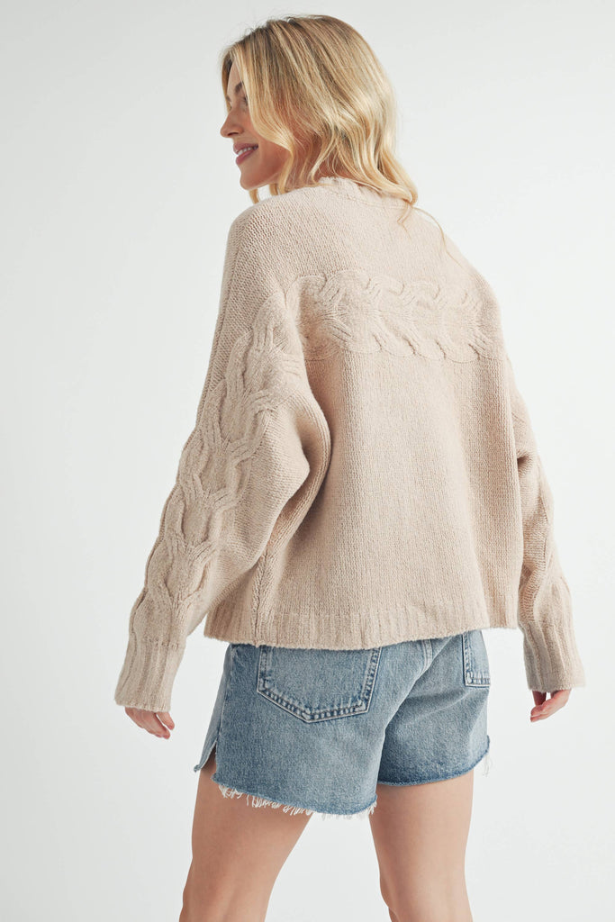 Woman wearing a beige cable-knit sweater with relaxed fit and cable details on the chest and sleeves.