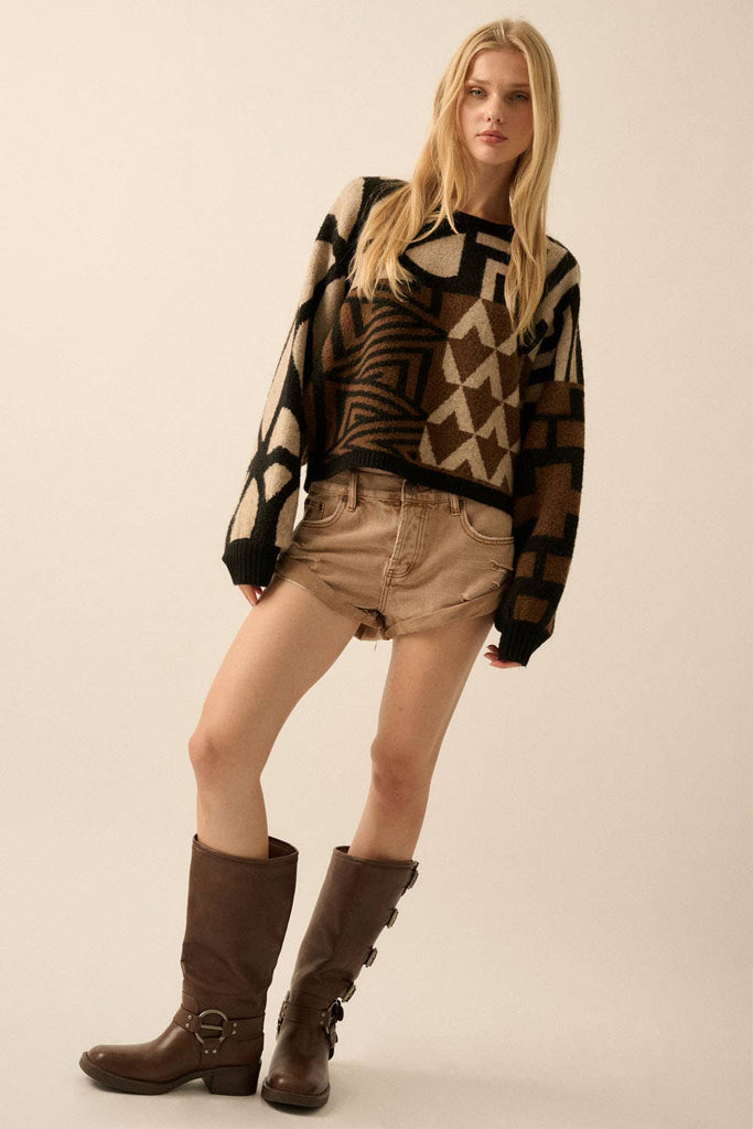 Model wearing a patterned knit sweater with bold geometric designs in neutral and contrasting tones, styled with casual bottoms.